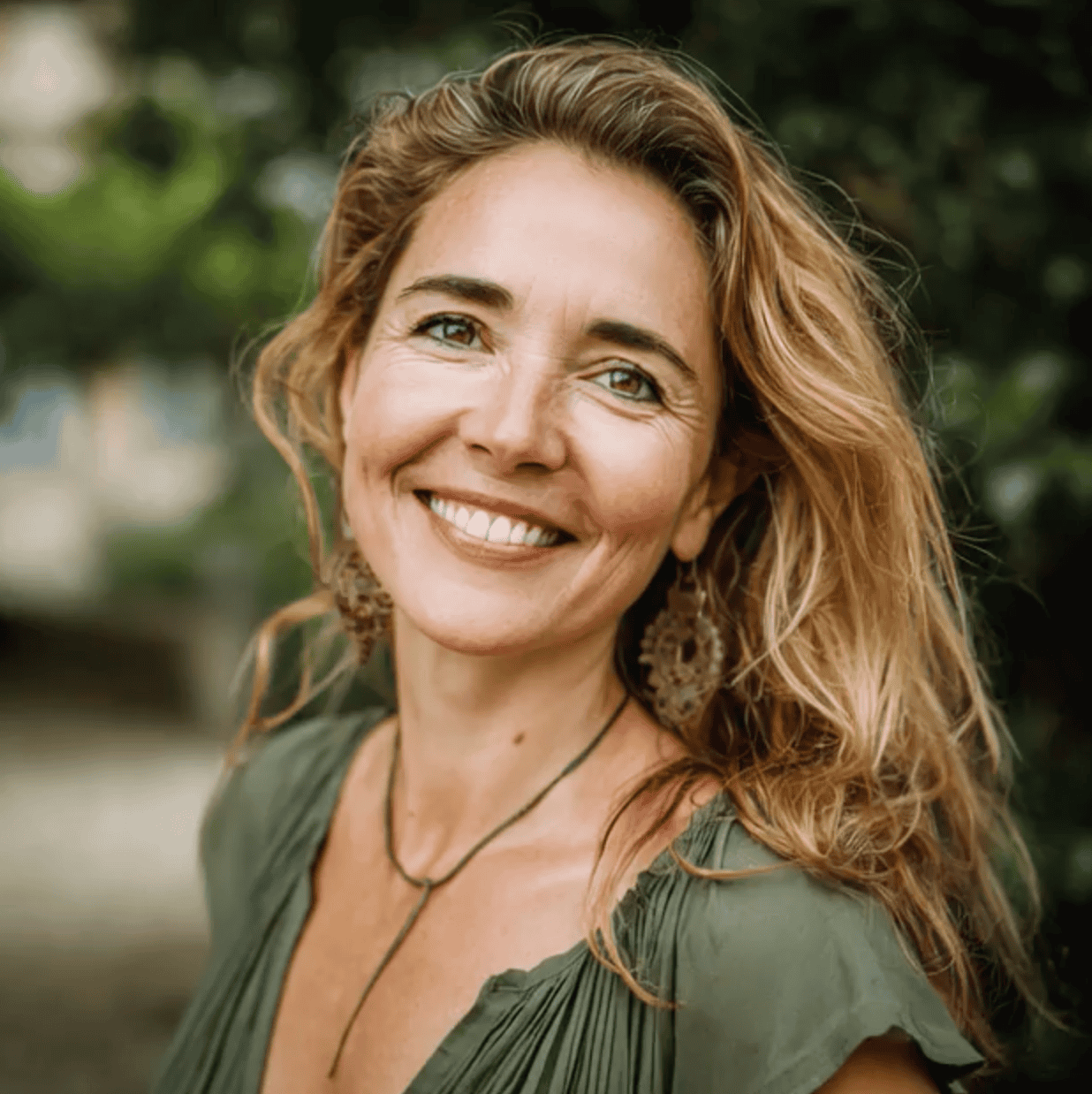 Smiling woman with wavy red hair wearing boho earrings and a green top outdoors, natural background, portrait shot for lifestyle or fashion SEO purposes.
